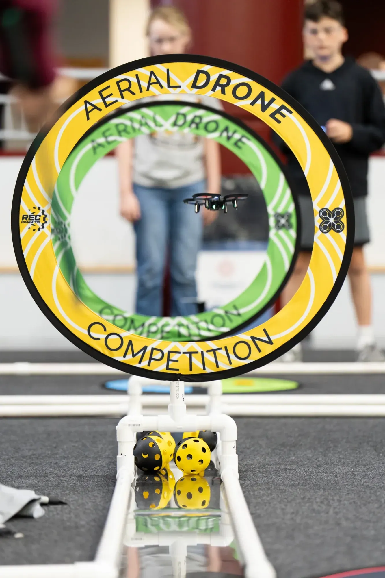 Un dron CoDrone EDU en vuelo dentro del aro amarillo y verde de la Aerial Drone Competition, con dos estudiantes pilotando al fondo de la cancha