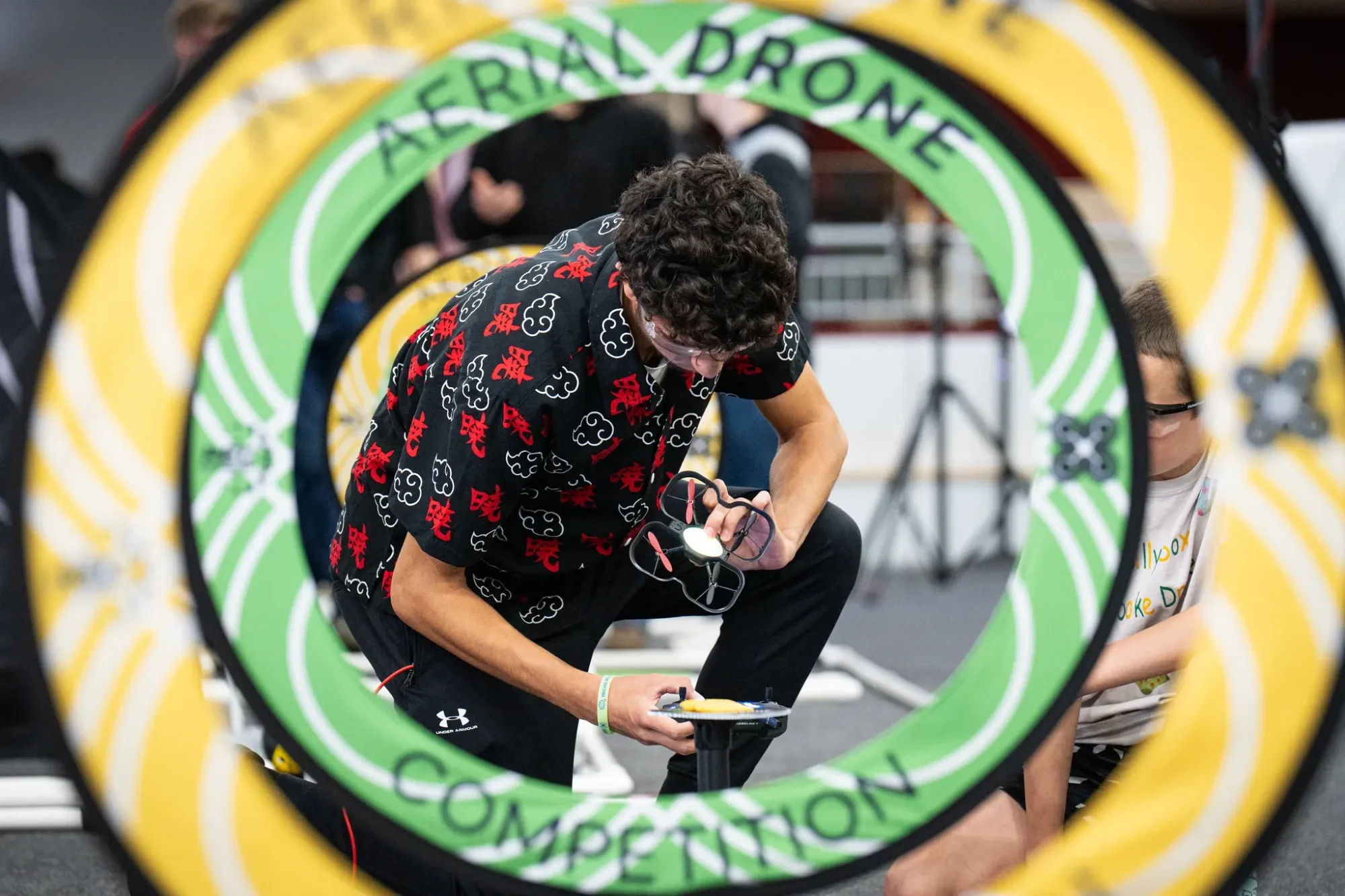 Un estudiante deja su dron sobre la plataforma de despegue, visto a través de un aro amarillo y verde de la cancha de la Aerial Drone Competition